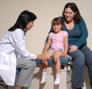 Healthcare provider using reflex mallet on girl's knee while woman looks on.