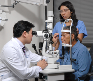 Ophthalmologist examining woman's eye with lens and slit lamp. Another healthcare provider holding patient's head steady.