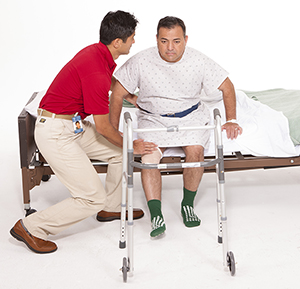 Healthcare provider helping man stand up from hospital bed with walker nearby.