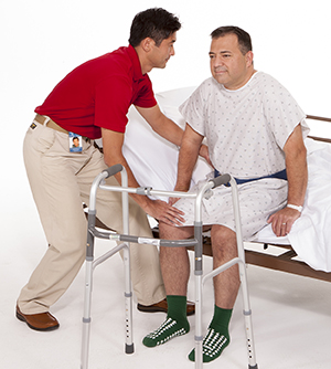 Proveedor de atención médica ayudando a un hombre a levantarse de la cama del hospital. Hay un andador al lado de la cama.
